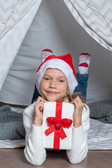 A girl in a Santa hat holds his big gift box for Christmas New Year lying in a children's tent wigwam in the nurser © Alena