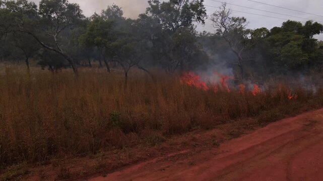 Aerial view of grassfire flaring up beside red dirt road in Cape York, Queensland, Australia. Long dry grass burns with bright flames and billowing white smoke in the remote Australian Outback. 45