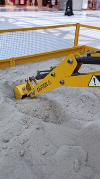 A close-up vertical shot of a yellow miniature toy excavator bucket digging and scooping loose sand. The mechanical arm operates inside a fenced indoor sandbox play area at a brightly lit shopping mal