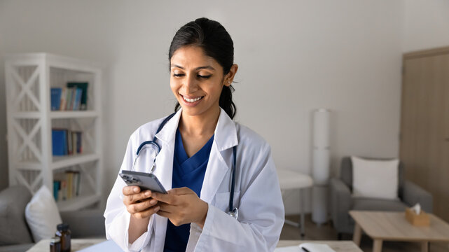 Smiling South Asian woman doctor texting on smartphone in clinic, checking information, replying on patient message, conduct medical consultation through telemedicine. Mobile application, connectivity