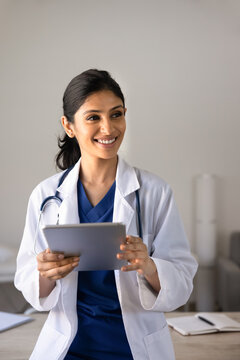 South Asian woman doctor smiling holding digital tablet in medical clinic, represents healthcare professional using technology, telemedicine application in patient care and online remote consultations