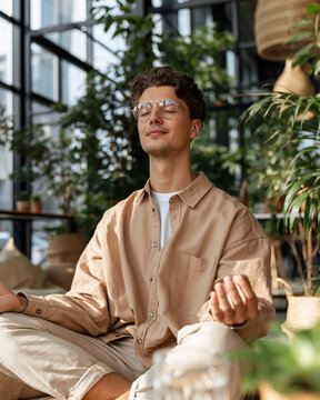 Young man meditates calmly in sunlit, plant-filled space. Glasses and light shirt frame his peaceful, closed-eyed expression. Fingers gently clasped, posture relaxed, suggesting inner stillness