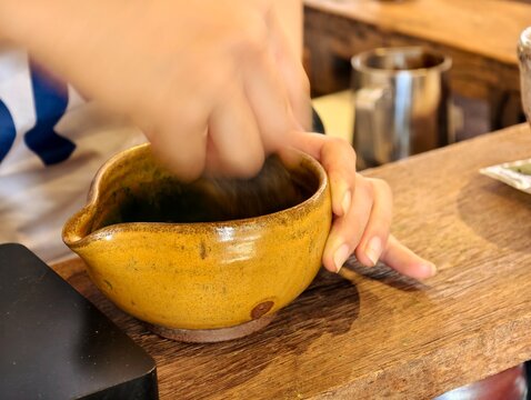 A detailed action close-up of a barista using a bamboo chasen whisk to prepare green tea matcha in a handcrafted yellow ceramic bowl. Features motion blur, symbolizing authentic, fresh preparation.