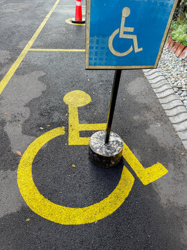 Yellow wheelchair symbol painted on wet asphalt. Blue sign with white wheelchair icon stands beside it. Symbol marks reserved parking for disabled individuals. Surface shows rain splashes