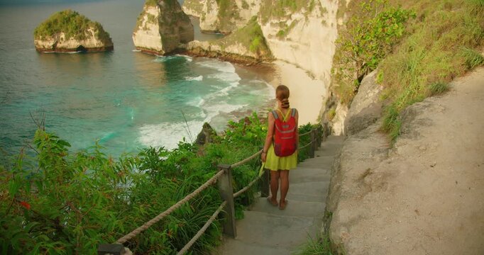 Tourist woman with backpack standing on stairs overlooking a blue bay and ocean in Nusa Penida