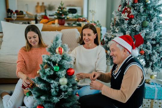 Adult daughter and senior parents enjoying Christmas tree decorating together at home, sharing happy family moments surrounded by ornaments and holiday gifts.