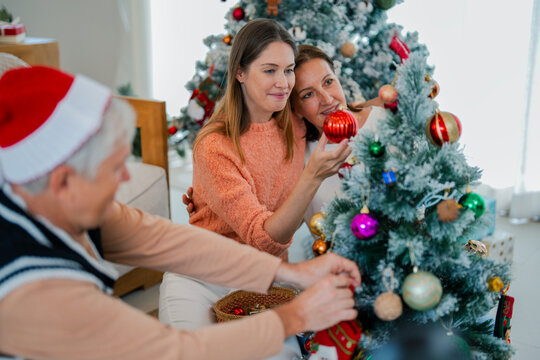 Senior mother embracing adult daughter while family decorates Christmas tree together, showing love, togetherness and joyful holiday tradition at home.