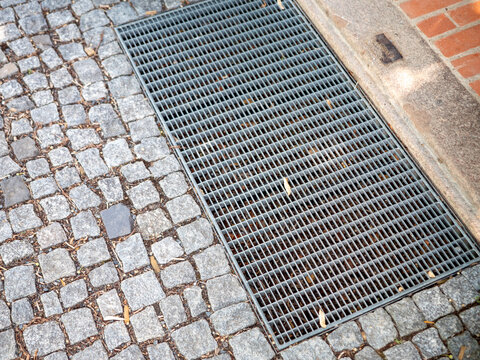 Metal drainage grate set in a cobblestone pavement at a building entrance, showing urban drainage, stone paving, grid texture and architectural detail.