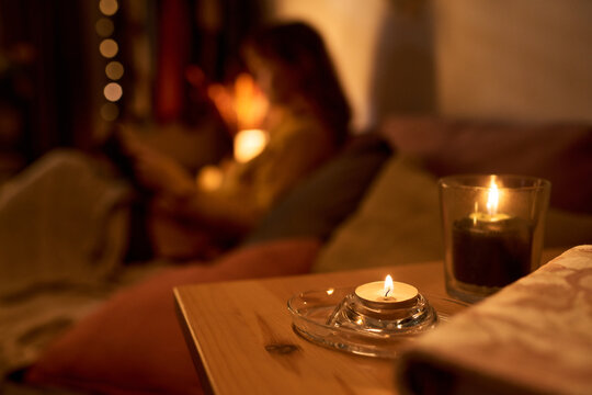 Lit candles on wooden table with blurred Caucasian young adult woman reading book on sofa in background, representing mental wellbeing and self care, close up shot