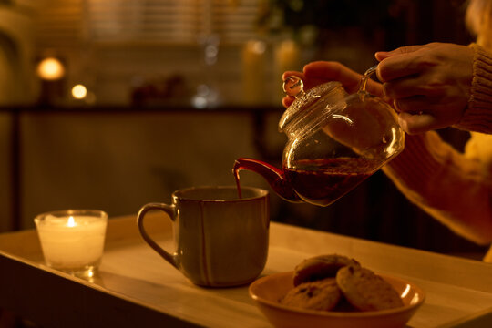 Unknown woman pouring hot tea from glass teapot into ceramic mug on tray with cookies. Image represents self care and relaxation, ideal for mental wellbeing campaigns
