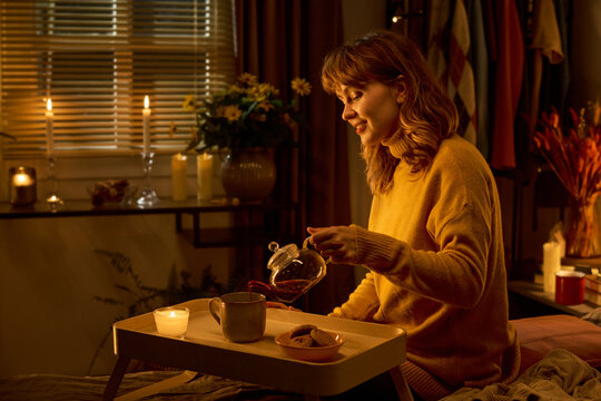 Young adult Caucasian woman pouring hot tea from glass teapot into mug on bed tray, practicing self care at home. Image suitable for mental wellbeing and cozy lifestyle marketing