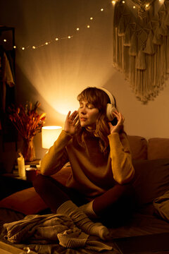 Young adult Caucasian woman sitting cross legged on bed wearing headphones with eyes closed, listening to music or meditating, representing mental wellbeing, vertical