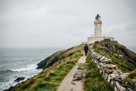 A person walking on a narrow path towards a white lighthouse on a cliff edge, with a stone wall and the sea visible under a grey, cloudy sky
