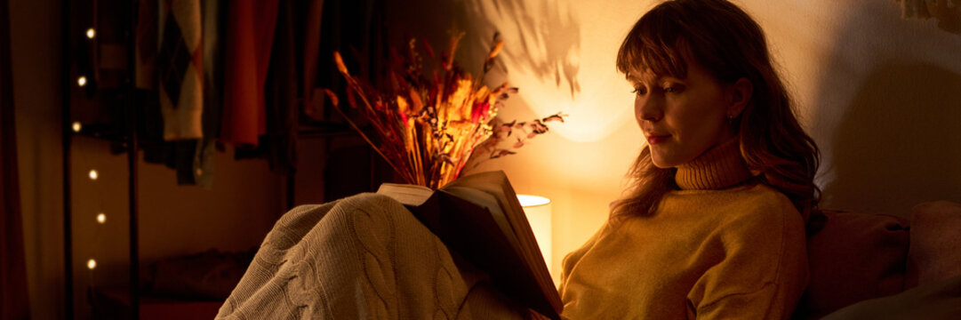 Caucasian young adult woman reading book in bed under warm lamp light, enjoying quiet evening. Image suitable for mental wellbeing campaigns and leisure lifestyle marketing