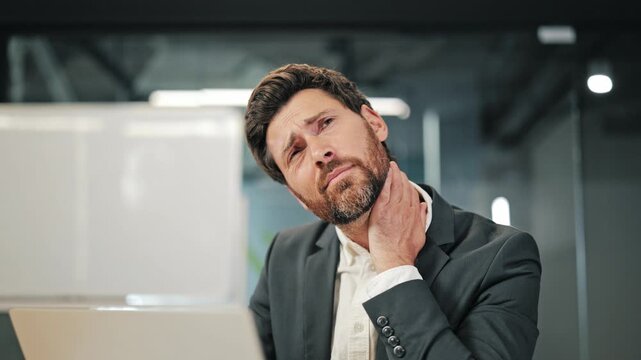Man experiences neck pain at office workstation. Discomfort evident while looking at laptop. Portrays work-related strain, showing need for ergonomic relief and stress management during long hours.