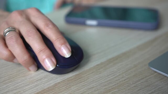 A businesswoman's hand working on a laptop, with a smartphone lying on a desk. Close-up of a computer mouse in her hand.