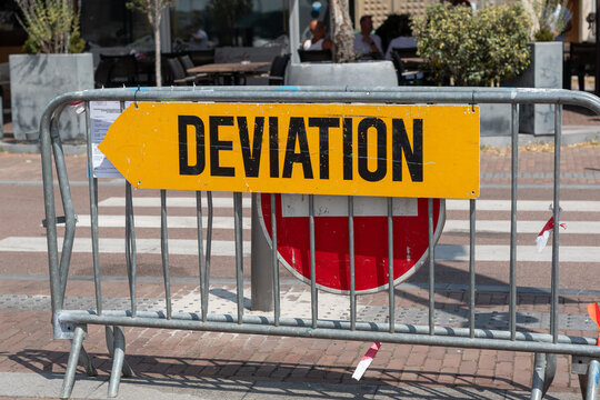 Yellow deviation traffic sign on a metal barrier block road with a no entry sign in the background.