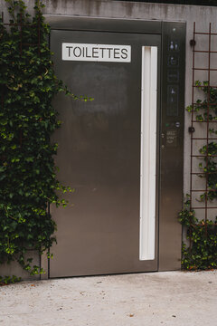 Modern stainless steel public toilet door with "Toilettes" sign in French. Automatic metal door of a public restroom surrounded by green climbing plants on an outdoor wall.