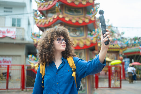 Caucasian woman travel vlogger filming selfie video at colorful Chinese temple shrine in Thailand with action camera