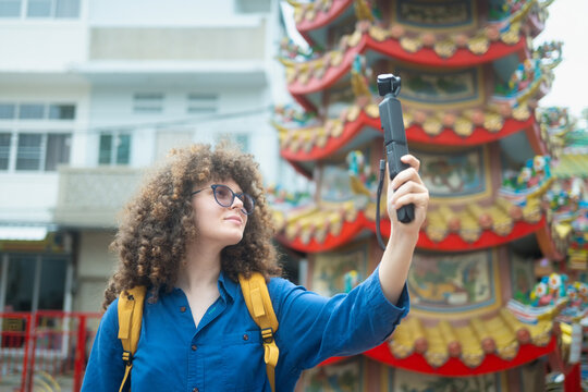 Caucasian woman travel vlogger filming selfie video at colorful Chinese temple shrine in Thailand with action camera