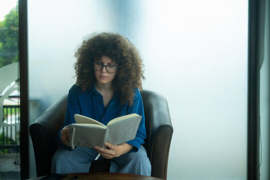 Caucasian Woman with Curly Hair Reading Book in Armchair by Window