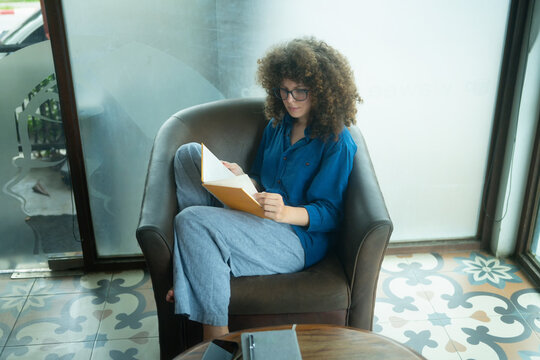 Caucasian Woman with Curly Hair Reading Book in Armchair at Cafe