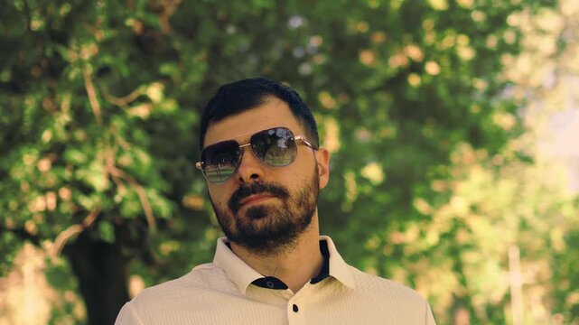 Various video clips of a bearded man wearing sunglasses and a polo shirt walking in a green park. He strolls along a paved path and adjusts his sleeve under natural daylight among trees.