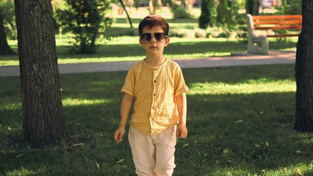 Cute little boy wearing sunglasses and a yellow shirt walking in a sunny green park during summer. Happy childhood and outdoor lifestyle portrait of a fashionable child enjoying nature.
