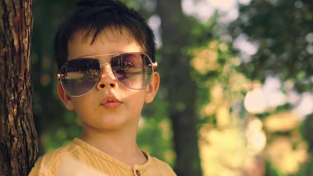 Cute little boy wearing sunglasses and a yellow shirt walking in a sunny green park during summer. Happy childhood and outdoor lifestyle portrait of a fashionable child enjoying nature.