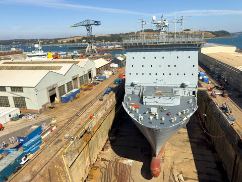 Falmouth Cornwall UK. 13.08.2026. A British navy grey ship is positioned in a deep dry dock, surrounded by industrial buildings and heavy machinery, indicating ongoing maritime construction