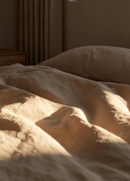 Close-up of a bed with a beige-colored bedsheet and a white pillow. the bed appears to be in a bedroom, as there is a window with sheer curtains in the background.