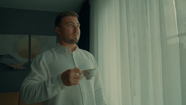 The man is standing in a shirt and drinking morning coffee by the window. The man is getting ready for serious work.