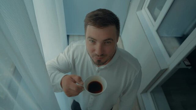 A man in a shirt is watching the frame and drinking morning coffee. A demonstration of drinking tea and other beverages. A bold expression on a handsome face.
