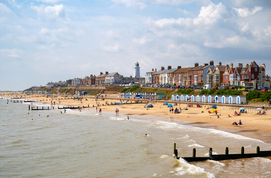 Colourful beach huts line the promenade in Southwold, Suffolk, with its iconic lighthouse rising above the town. Families relax and swim on the sandy beach under a bright summer sky.