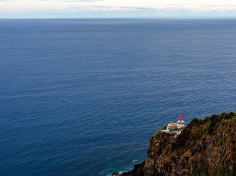 Farol do Arnel lighthouse on Nordeste cliffs. Perched on a steep volcanic cliff. Vast Atlantic Ocean to the horizon. Sao Miguel, Azores coast. Portugal.