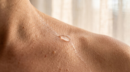Fototapeta premium Macro close up showing single clear water droplet running down smooth human female shoulder bare skin illuminated by bright natural morning indoor sunlight