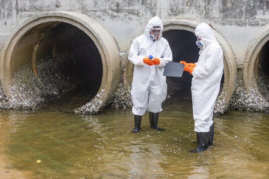 scientists in hazmat suits with tablet inspecting wastewater drainage pipe, environmental researchers in protective gear checking water pollution by tunnel
