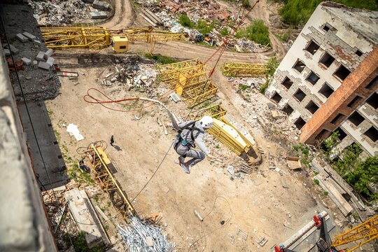 Aerial view of person in helmet rope jumping with arms spread over demolition site near abandoned building, extreme sport adrenaline and thrill concept