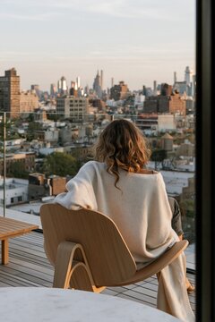 Young woman in oversized cardigan and slim jeans seated in curved teak chair on urban rooftop terrace with skyline view during golden hour