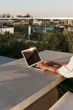 Woman's hands typing on a laptop on a low concrete table, urban rooftop setting with warm sunset light illuminating the scene