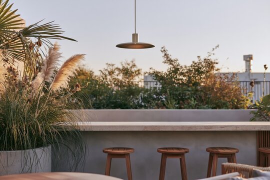 Urban rooftop bar corner featuring a brushed brass pendant light over a low travertine counter with teak stools and pampas grass in a concrete planter