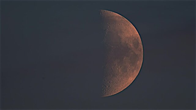 A half moon hangs in the dark night sky. The lunar surface displays craters and textured terrain in shades of orange and grey against the deep blue atmosphere.