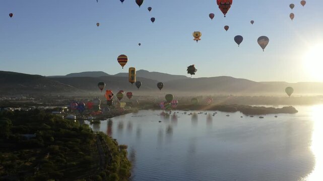 Cinematic wide aerial drone shot of dozens of colorful hot air balloons floating over a calm lake at sunrise, golden light reflecting on the water surface,mountains in the background,soft morning mist
