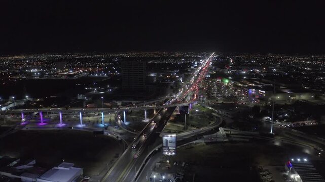 Nighttime aerial drone shot flying over a modern highway overpass interchange with colorful glowing purple and blue LED lights, light trails from moving traffic, illuminated city skyline.