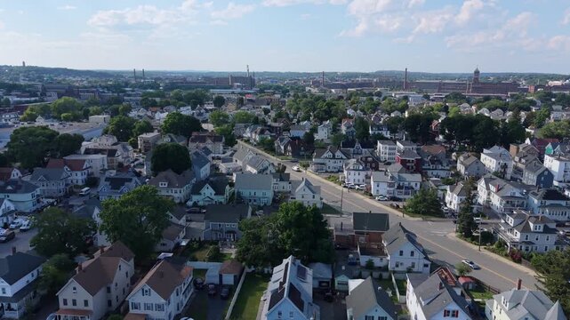 Dense rows of multi-family houses with gabled roofs and porches line narrow streets, cars fill driveways while historic mill buildings rise above Lawrence, Massachusetts.Aerial drone lateral wide shot