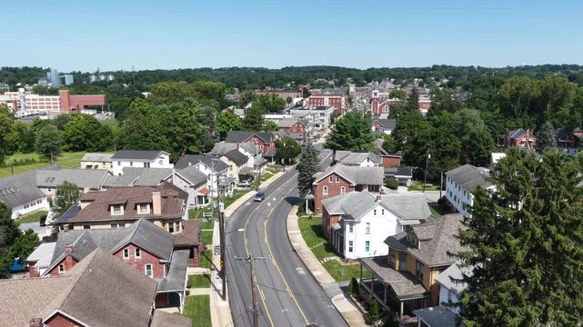Curving street winds past historic brick and white houses with tall pine trees, single car drives toward distant brick factory buildings in Elizabethtown, Pennsylvania. Aerial drone wide shot