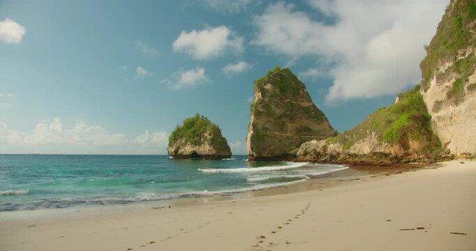 Footprints lead across pristine white sand toward large rock formations in blue ocean water
