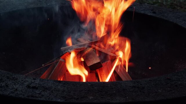 Wood burning inside an outdoor fire pit with bright orange flames, smoke and small sparks. Close-up view of a warm campfire burning in the evening.