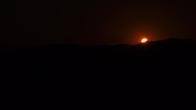 Partial solar eclipse setting behind a dark mountain ridge at sunset. Bright orange sun slowly disappears below the horizon under a deep red evening sky.