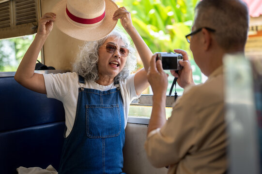 Asian senior elderly woman posing playfully with sun hat while her partner takes a photo with camera